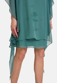 Abito in chiffon turchese con design a strati, caratterizzato da un orlo asimmetrico, una struttura leggera e tessuto drappeggiato. Accentuato da maniche fluttuanti.