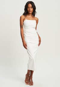 White, sleeveless midi dress with ruched detailing, made from a smooth fabric. Model wears clear heeled sandals with a simple strap design.