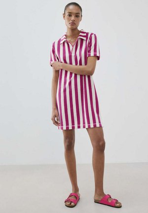 Frau steht, trägt ein kurzärmliges, kragenbehaftetes Kleid mit vertikalen rosa und weißen Streifen und rosa offene Zehensandalen vor einfarbigem Hintergrund.