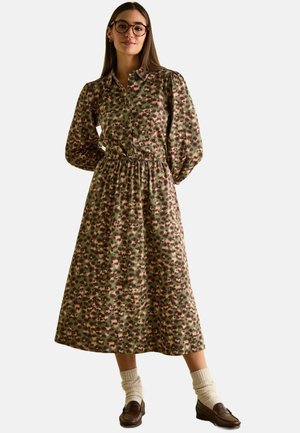 Grünes Midi-Kleid mit Blumenmuster, langen Puffärmeln, Knopfkragen und elastischem Taillenbund. Getragen mit cremefarbenen Socken und braunen Slippern.