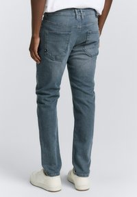 Mann in hellblauen geraden Jeans und weißen Sneakers, der mit hängenden Händen vor einem schlichten Hintergrund steht.