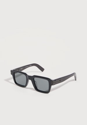 Black rectangular sunglasses with dark tinted lenses and thick arms, resting on a light gray surface.