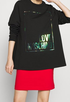Modèle portant un haut noir à manches longues avec l’inscription holographique Love Moschino et une jupe rouge vif.