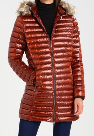 Femme portant un manteau matelassé brillant couleur rouille orangé avec une capuche bordée de fourrure, une fermeture éclair noire et des poches latérales, associée à un pantalon bleu foncé.