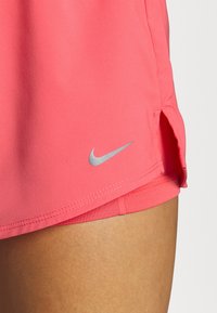 Rosa sportshorts med en silverfärgad Nike-logotyp. Har sidoventiler och en lätt, slät textur. Designen inkluderar en kontrasterande innerfoder.