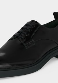 Chaussure habillée en cuir noir poli à lacets avec bout arrondi et étiquette de marque sur le côté près des lacets.