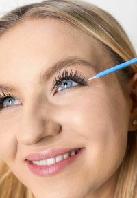 Blå applicatorstav placerad nära ögat, som förstärker långa, mörka ögonfransar. Motivet har ljus hy, blont hår och ett subtilt leende.