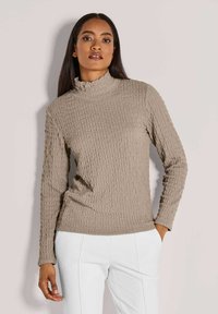 Frau in einem strukturierten beigen Rollkragenpulli und weißen Hosen, die mit einer Hand in der Tasche vor einem hellen Hintergrund steht.