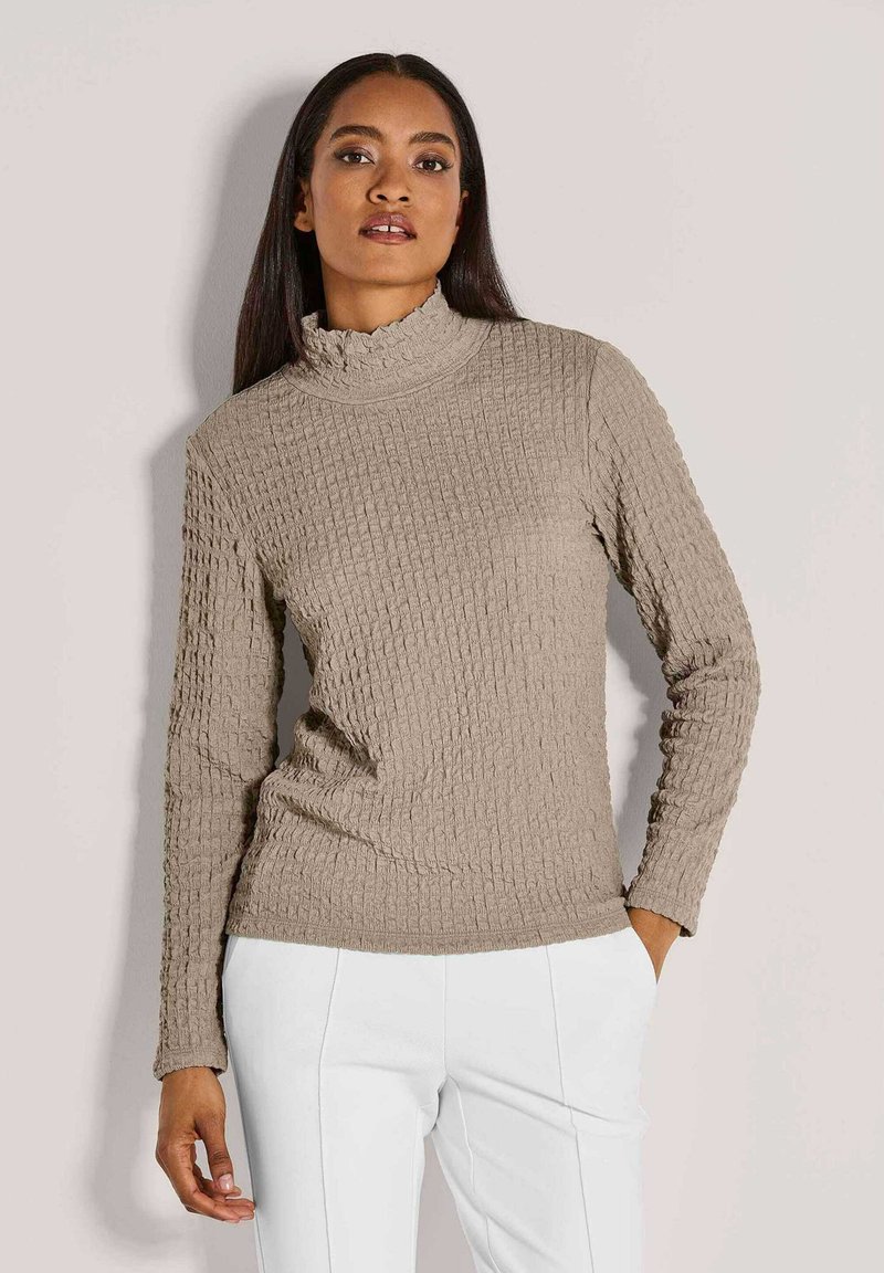 Frau in einem strukturierten beigen Rollkragenpulli und weißen Hosen, die mit einer Hand in der Tasche vor einem hellen Hintergrund steht.