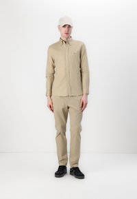 Beige gombos ing és világosbarna nadrág, fehér sapkával és fekete cipővel párosítva. Egyszerű dizájn, látható minták vagy díszítések nélkül.
