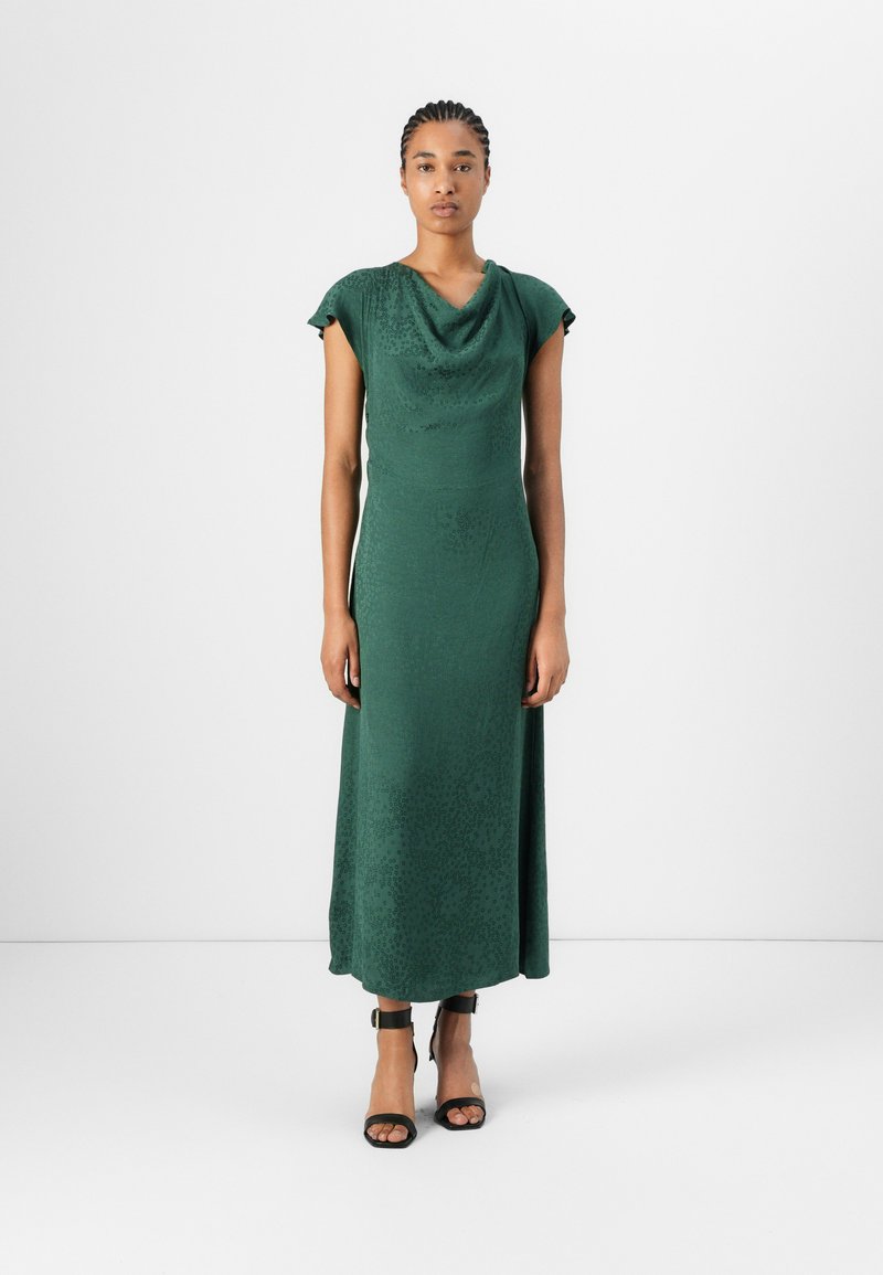 Green, mid-length dress with a cowl neckline and short sleeves. Made of a textured fabric featuring a subtle pattern. Worn with black strappy sandals.