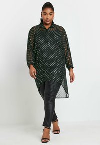 Blusa nera trasparente con bottoni e pois bianchi, maniche lunghe, orlo a strati e alta-bassa, indossata sopra leggings in pelle nera e tacchi con cinturini.