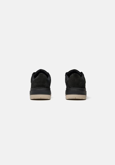 Paire de baskets basses noires vues de dos, avec des semelles beiges et des motifs croisés cousus sur les talons.