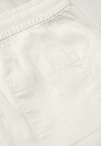 Gros plan sur un tissu beige clair avec une ceinture élastique et une poche arrière rectangulaire cousue sur un pantalon décontracté.