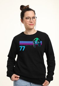 Mujer con gafas y moño, vestida con sudadera negra con un gráfico colorido de TIE Fighter, rayas azules y el número 77, de pie contra un fondo blanco.