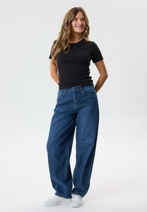 Mujer con cabello largo y ondulado, vestida con una camiseta negra de manga corta, jeans azules holgados y zapatillas blancas, de pie con las manos detrás de la espalda.