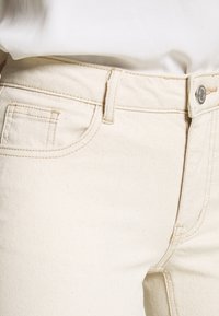 Pantalones vaqueros de mezclilla beige con bolsillos frontales, cierre de botón de metal y detalles de costura visibles. Textura suave con un sutil patrón de grano.