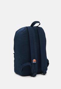 Zaino di tessuto blu navy con spallacci regolabili, maniglia superiore e un piccolo patch logo sul lato. Texture liscia e forma arrotondata.