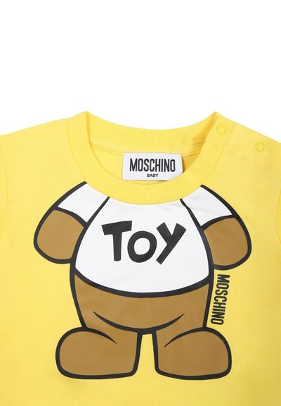 T-shirt di cotone gialla con grafica di un orso e la scritta "TOY" stampata su uno sfondo bianco. L'etichetta del brand "Moschino Baby" è visibile.