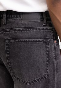 Μαύρο τζιν παντελόνι με λεία υφή, διακοσμημένο με μια μεγάλη πίσω τσέπη και μια ορθογώνια δερμάτινη ετικέτα στη ζώνη μέσης.