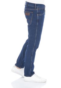 Niebieskie dżinsy o wąskim kroju z prostą nogawką, klasycznym wzorem z pięcioma kieszeniami i kontrastowym szwem wzdłuż bocznych szwów. Widoczne białe sneakersy.