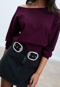 Pull bordeaux à épaules dénudées avec des poignets côtelés, associé à une mini-jupe en jean noire et une large ceinture noire avec des doubles boucles argentées.