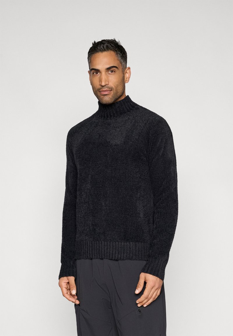 Schwarzer, strukturierter Strickpullover mit hohem Kragen und geripptem Saum. Schmale Passform, lange Ärmel und weiche Stoffstruktur. Kombiniert mit dunkler Hose.