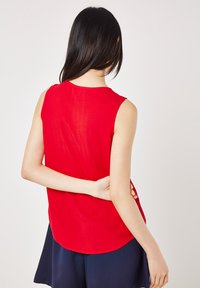 Red sleeveless top featuring a textured pattern with a curved hem and a casual fit, paired with dark shorts.