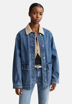 Jeune femme portant une veste en jean bleue surdimensionnée avec col beige, cardigan beige, haut noir, jean bleu clair et ceinture blanche sur fond blanc.