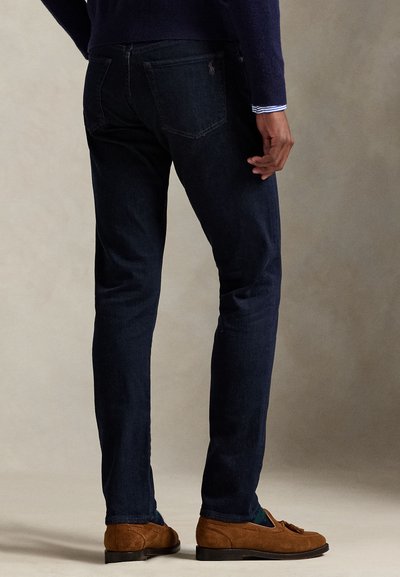 Mørkeblå, tettsittende jeans med en glatt tekstur og subtile søm detaljer. Brukt med brune semskede loafers. Vist bakfra.