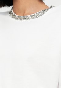 Λευκό μπλουζάκι με στρογγυλή λαιμόκοψη διακοσμημένη με διάφορες διάφανες στρας σε διαφορετικά σχήματα και μεγέθη, προσθέτοντας υφή και λεπτομέρεια.