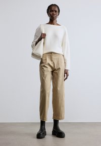 Fehér kötött pulóver, bézs nadrág, és fekete bokacsizma. Modell fehér táskát tart. Egyszerű kialakítás, laza szabás. Simogatott textúra.