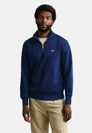 Man met baard die een marineblauwe kwart-ritssluiting trui draagt over een wit overhemd en beige broek, staand tegen een witte achtergrond.