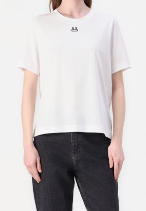 Kobieta ubrana w prostą, białą koszulkę z krótkim rękawem, ozdobioną małą czarną haftowaną twarzą i sercami przy dekolcie, w połączeniu z czarnymi dżinsami.