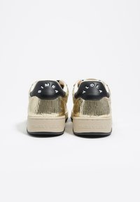 Zapatillas deportivas con una parte superior dorada agrietada, capuchón negro en el talón con marca blanca, suelas beige suaves y un acabado texturizado en la parte trasera.