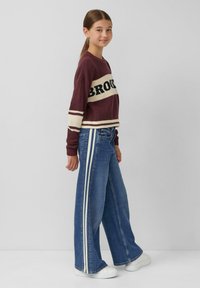 Maglione a coste color granata con "BROOKS" in nero, abbinato a jeans blu a gamba larga con strisce laterali bianche e sneakers bianche.