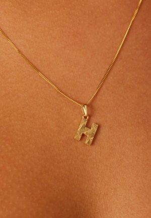 Collier en or avec un pendentif lettre "H" texturé, doté d'une chaîne fine et d'une finition polie, reposant sur une surface couleur peau.