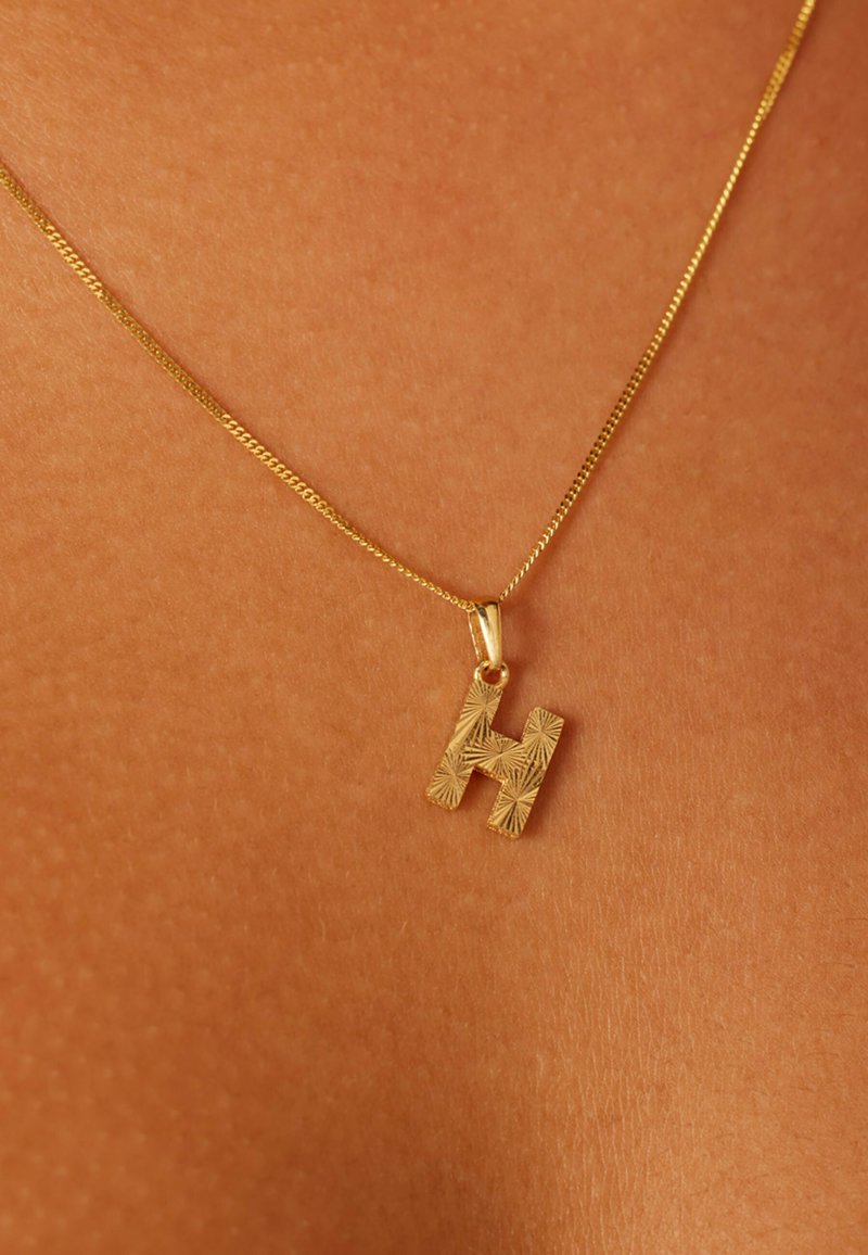 Gold necklace with a textured letter "H" pendant, featuring a thin chain and a polished finish, resting on a skin-toned surface.