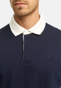 Granatowa koszulka polo z białym kołnierzykiem, krótkim zapięciem na guziki i haftowanym logo z rokiem "1993". Wykonana z tkaniny bawełnianej o teksturowanej fakturze.