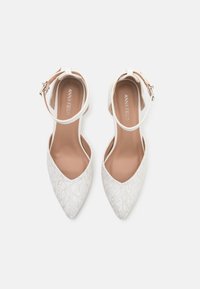 Scarpe bianche con cinturino alla caviglia in pizzo, con design a punta, caratterizzate da una superficie testurizzata e una soletta liscia di un leggero colore beige.