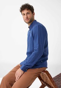 Camicia blu a maniche lunghe con colletto con bottoni, tessuto texturizzato e tasca frontale; abbinata a pantaloni marroni, seduto su una sedia di legno.