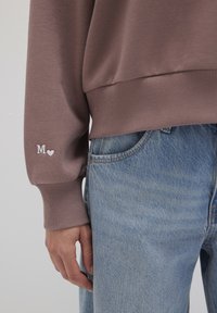 Light brown sweatshirt with ribbed cuffs, featuring a white embroidered "M" and heart. Paired with faded blue denim jeans.