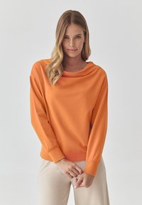 Oranžová dlouhá halenka s lodním výstřihem, volný střih a žebrovanými manžetami, kombinovaná se světlými béžovými kalhotami. Hladká textilie.