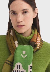 Pull vert en tricot associé à une écharpe à motifs représentant des maisons et une fleur. L'écharpe présente des tons terreux et des accents géométriques.