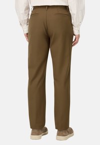 Pantaloni marroni con una vestibilità su misura, dotati di due tasche posteriori. Abbinati a una camicia di colore chiaro e sneaker beige. Texture del tessuto liscia.