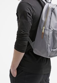 Šedý látkový batoh se zlatým zipem a hnědým koženým akcentem, nošený na zádech osoby v černém tričku s dlouhým rukávem.