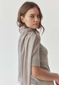 Écharpe gris clair en tissu doux, drapée élégamment autour de l'épaule. Présente un léger détail à frange aux extrémités.