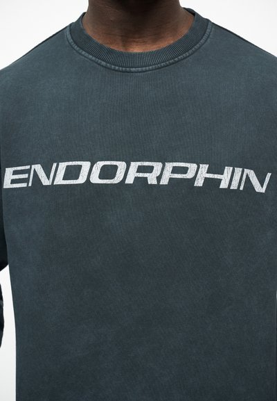 Gros plan sur une personne portant un t-shirt gris foncé à col rond avec le texte blanc "ENDORPHIN" en gras, police légèrement usée, sur la poitrine.