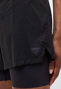 Μαύσες αθλητικές βερμούδες με διακριτικό λογότυπο πάνθηρα στο τελείωμα, φορεμένες με αντίστοιχες μαύρες συμπιεστικές βερμούδες από κάτω στον μηρό ενός ατόμου.