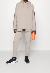 Beżowa bluza z kapturem z czarnymi paskami po bokach i kieszenią z przodu, zestawiona z dopasowanymi zwężanymi spodniami. Czarne sneakersy i pomarańczowa butelka na wodę.